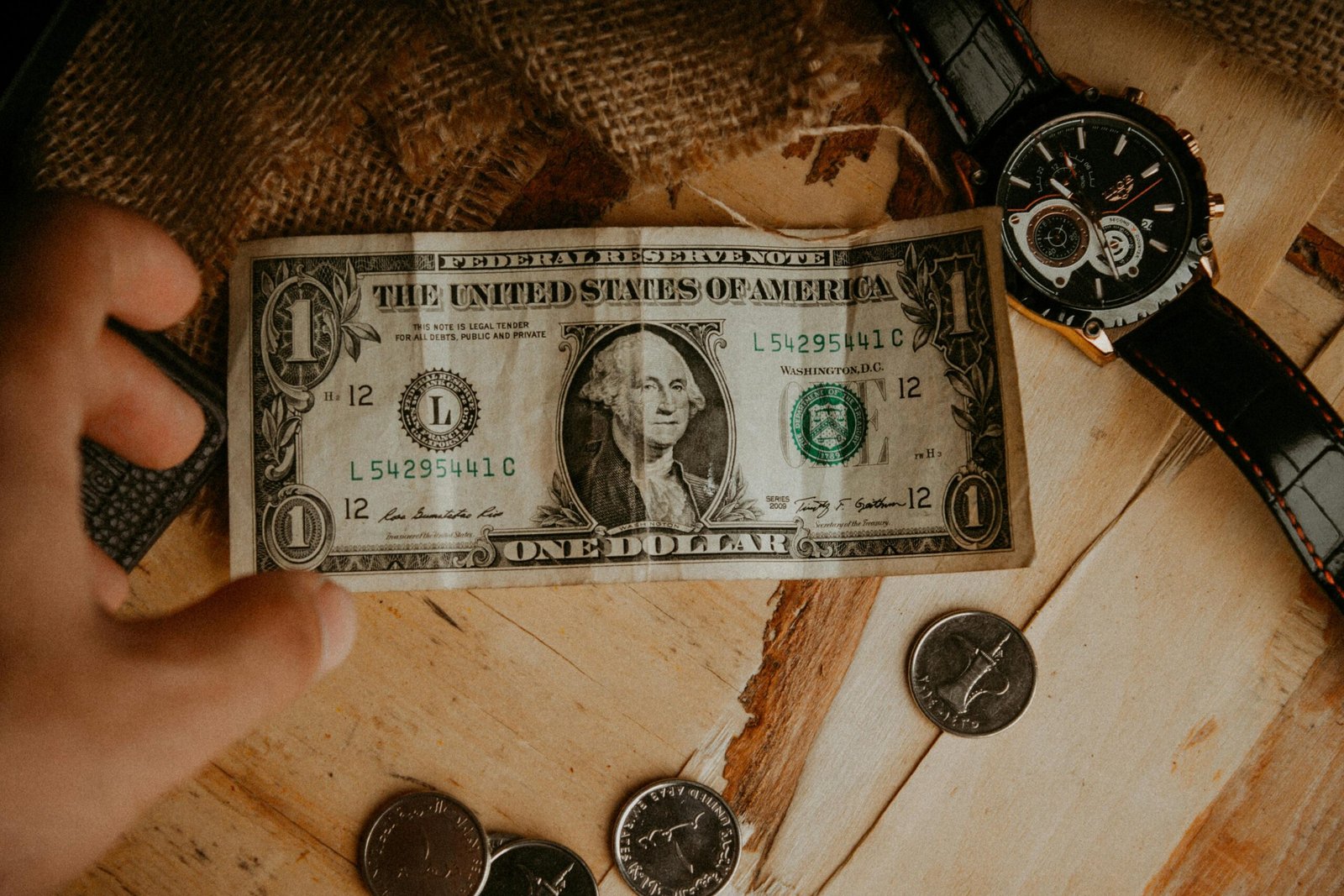 A person reaches for a one dollar bill surrounded by coins and a watch on a wooden table.