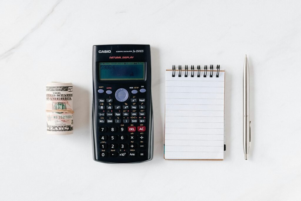 From above of roll of American dollar banknotes tied with rubber band near professional calculator and empty spring notepad with pen on marble table