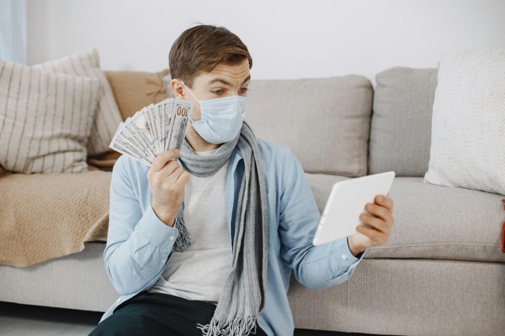 A man in a face mask holding cash and using a tablet in a cozy living room setting.