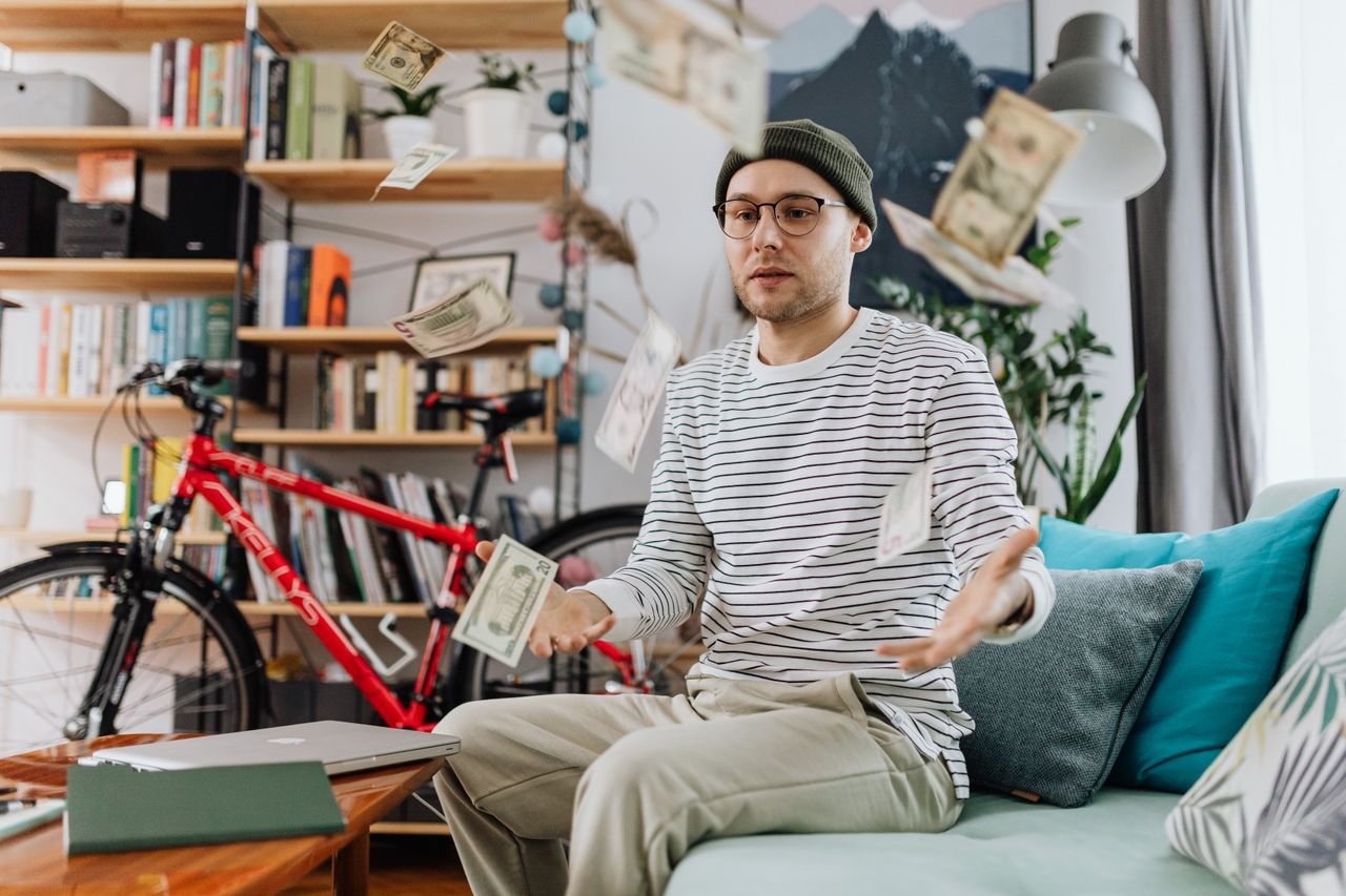 Young man tossing money in a cozy room, showcasing indoor leisure and wealth.